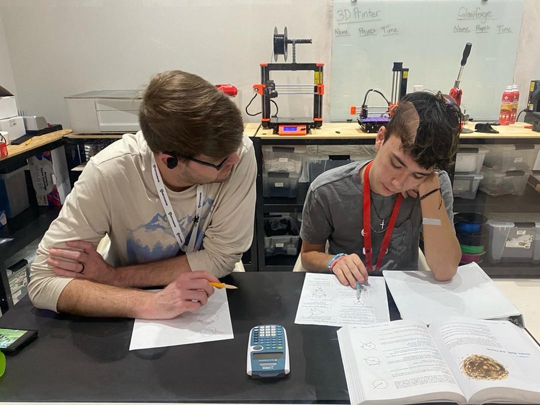 A guide helping a learner with math at a table in front of the 3D printers